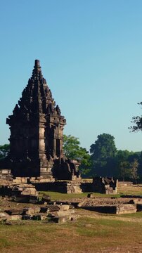 A solitary ancient shrine stands among weathered archaeological ruins and dry grass within the expansive Prambanan Temple complex.