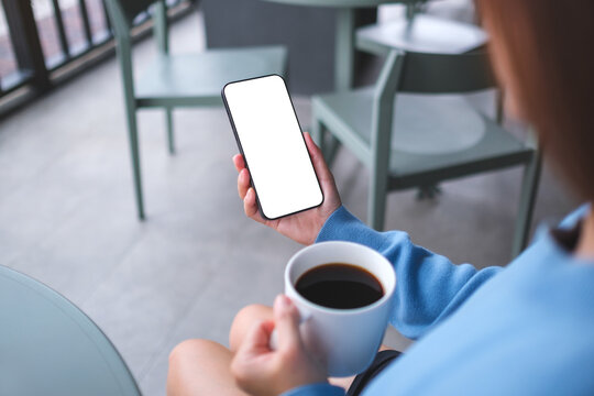 Mockup image of a woman holding and using mobile phone with blank desktop screen while drinking coffee in cafe