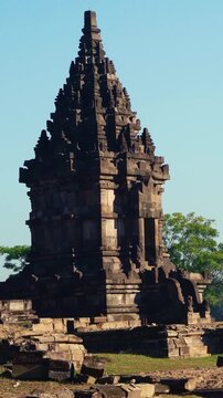 Historic Prambanan shrine rises above scattered stone foundations and archaeological remains beneath a vivid tropical blue sky.