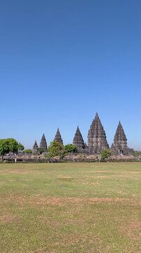 Distant Prambanan Temple spires rise across a broad grassy field beneath a clear blue tropical sky in Indonesia.
