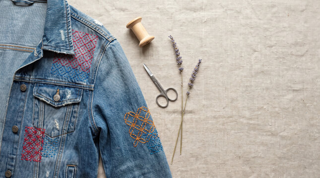 Embroidered denim jacket with colorful stitching, scissors, spool, and lavender beige fabric, showcasing creative textile
