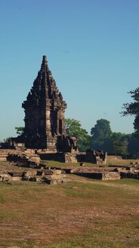 A small Prambanan shrine stands among ancient stone ruins and dry archaeological grounds beneath a clear blue sky.