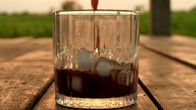 Pouring Dark Coffee into Glass with Ice Cubes on Wooden Table Outdoors