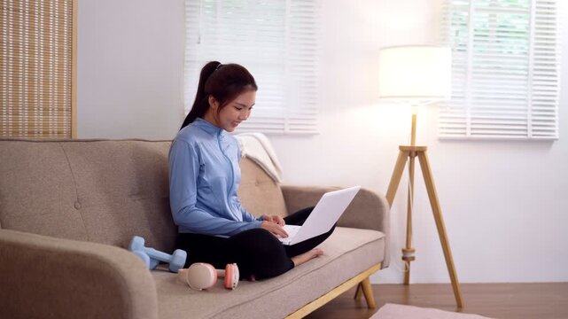 Young Asian woman practicing yoga with laptop on sofa next to dumbbells in bright living room