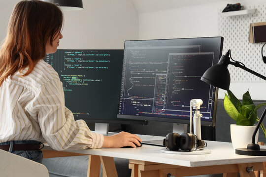 Female software developer coding on a desktop computer with multiple screens in a modern home office setting, the woman is focused on her work, using a mouse and keyboard