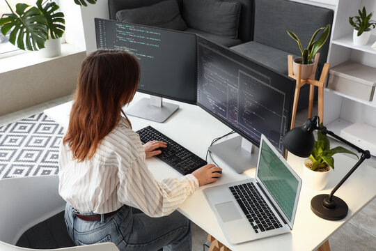 Female software developer coding on multiple computer screens and laptop in a modern home office environment, the modern home office environment supports remote work and digital innovation