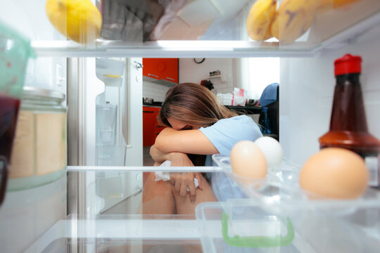 Sad Crying Woman Leaning On Open Refrigerator Expressing Emotional Eating Concept. Depressed lady weeps near the fridge door seeking comfort food through sadness
