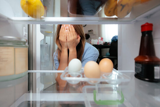 Sad Woman Covering Her Face Looking At Empty Fridge Inside Kitchen. Depressed female covers her eyes feeling upset near the poorly stocked refrigerator