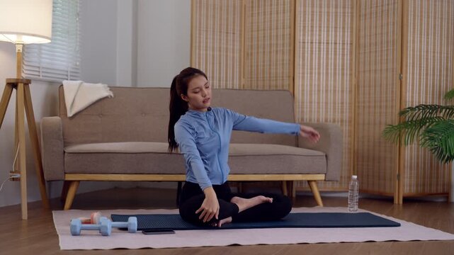 Young Asian woman performing seated side bend stretch on yoga mat during home fitness routine in modern living room