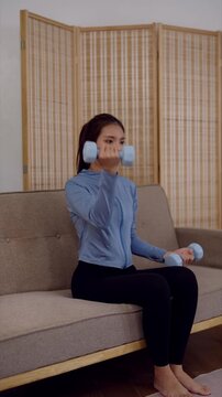 Young Asian woman performing bicep curls with dumbbells on sofa during home strength training session