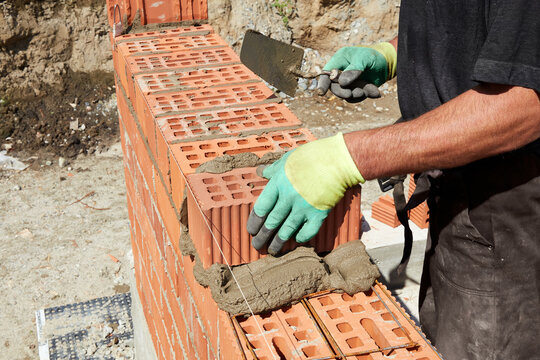 A skilled bricklayer is carefully applying mortar to red bricks, aligning them precisely to build a wall at a construction site