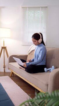 Young Asian woman wearing headphones reviewing paperwork on sofa during home fitness break in minimalist living room