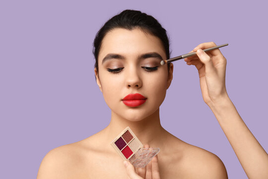 Female makeup artist working with young woman on lilac background