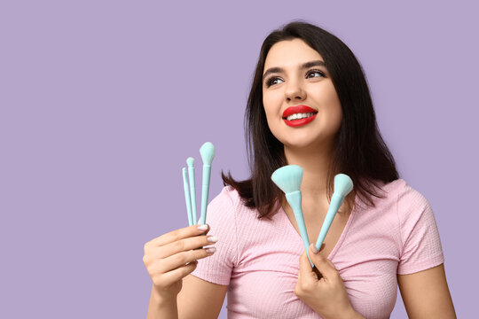 Beautiful young woman with makeup brushes on lilac background