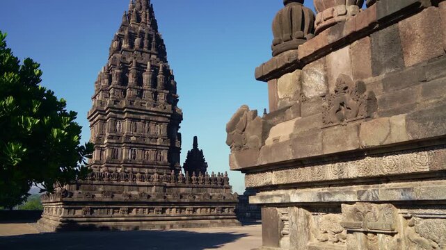 Sunlit courtyard view reveals a towering Prambanan shrine beyond ancient stone walls and shaded temple architecture.