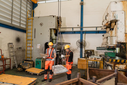 Worker holds a laptop while a colleague steadies a chain-suspended wrapped load, both standing between two heavy old power press machines