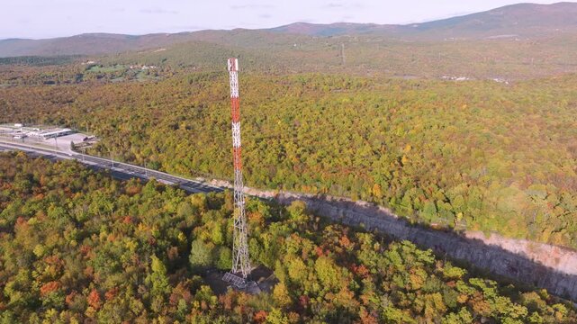AERIAL: Telecom tower rising above colorful sunlit autumn woodland and asphalt highway. Winding motorway winding through the hilly, forested landscape below, coloured in warm hues of fall season.