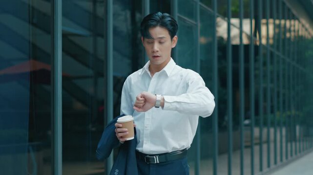 Asian businessman running late for work while checking wrist watch, holding coffee cup outside office building,representing hurry time management,man at work,work life business and wearable technology