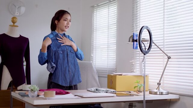 Young Asian fashion seller presenting blue denim shirt to camera while filming product showcase for online store