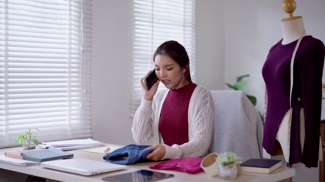 Young Asian fashion designer talking on phone while checking fabric samples at studio workspace