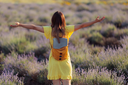  Woman In Yellow Dress And Backpack Standing With Open Arms In Lavender Field. Traveler seen from behind enjoys freedom inside a beautiful purple meadow