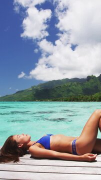 Luxury travel vacation destination bikini woman sunbathing on overwater bungalow deck at Tahiti resort tropical holiday. Raw unedited vertical video of real unretouched woman model without any makeup