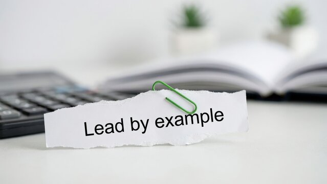 Torn paper note with Lead by Example text and green paperclip on an office desk