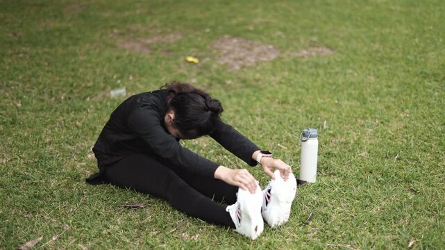High angle view of young Asian woman in black activewear sitting on grass, twisting torso and reaching to stretch hamstrings during outdoor warm-up in park.