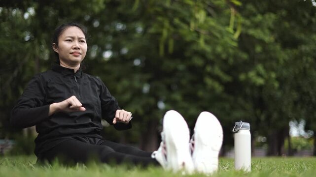 Asian young woman in sportswear sitting on park grass, stretching legs and reaching for toes for warm-up exercise near water bottle before outdoor running workout.