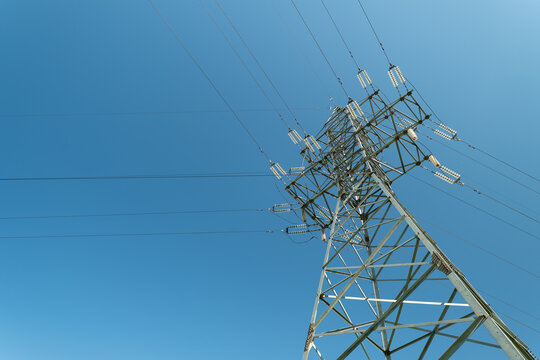 High voltage transmission tower rising above open landscape with power lines stretching across clear blue sky, seen from low angle with strong diagonal perspective and bright sunlight