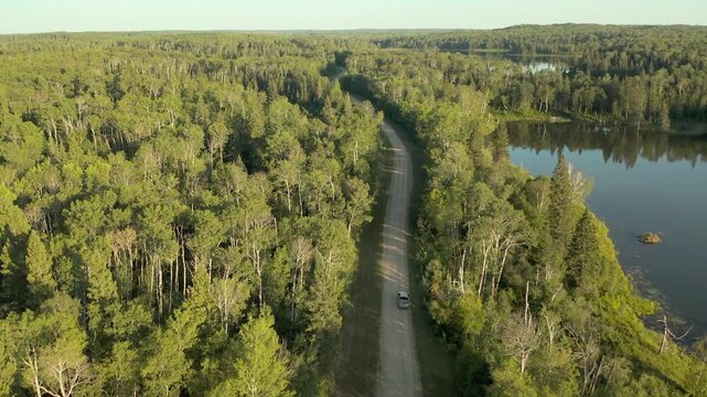 Aerial view of car driving on forest road, scenic road trip through lush green woodland, cinematic drone shot of vehicle traveling by lake