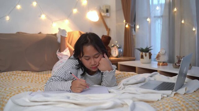 Exhausted young girl fell asleep while doing her homework in bed with notebook and laptop in warmly lit bedroom