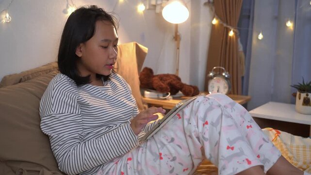 Young girl wearing comfortable pajamas using tablet while relaxing on bed in cozy bedroom at night