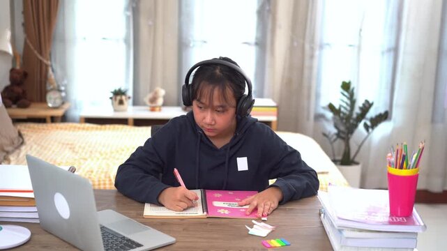 Young girl is wearing headphones and diligently taking notes on key points from her online lesson at home