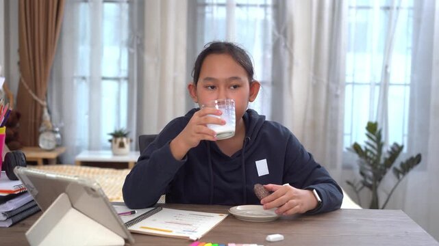 Happy young girl takes milk break while studying online at desk by notebooks and school supplies in home learning