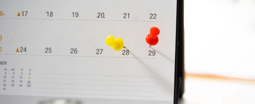 Yellow and red pins mark key dates on a calendar page. Soft light casts gentle shadows across the surface. The focus highlights small details like pin texture and date numbers. It suggests planning