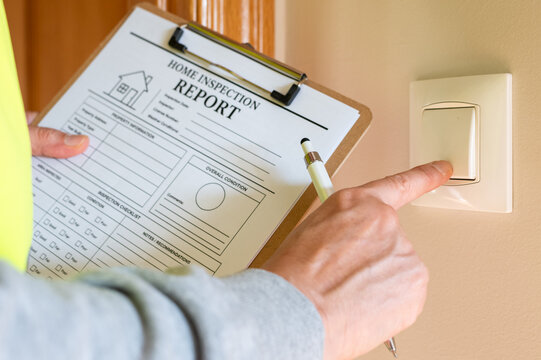 Professional home inspector documenting property condition and safety issues, examining an electrical light switch while holding a clipboard with a home inspection report form