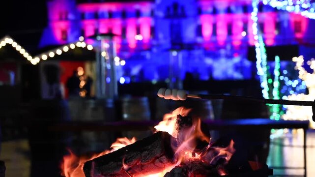 Person roasting marshmallows over burning bonfire at magical christmas market with illuminated castle background.
