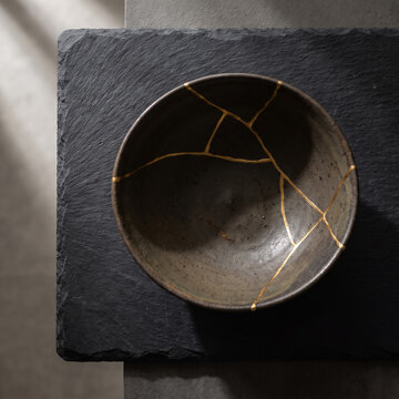Broken ceramic bowl mended with gold lacquer kintsugi technique on slate background minimalist dark fine art concept for resilience healing recovery philosophy