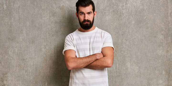 A serious man with a beard, wearing a white striped t-shirt, stands with his arms crossed against a gray textured wall. His expression is stern and unamused
