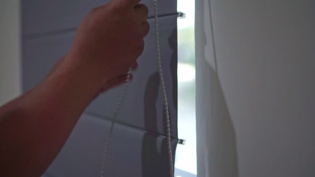 Human hand operating a window curtain chain to allow sunlight indoors. Residential lifestyle concept featuring privacy control and natural illumination.