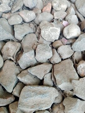 Gray stones scattered in rough, uneven piles. Some stones show faint pinkish highlights. Surface texture appears weathered, natural, rugged. Ideal for garden, landscaping, or construction projects