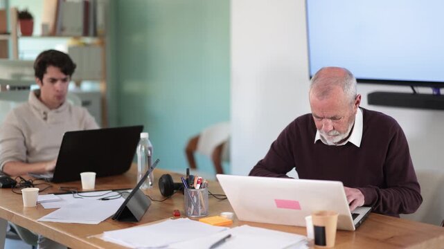  Elderly and young men colleagues working at desk using laptop in office 