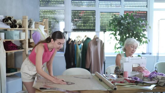  Elderly woman seamstress sews on machine adult woman assistant drawing pattern on paper in workshop 