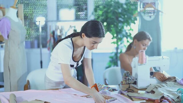 Team of dressmakers works in a sewing workshop. Fashion designer carefully cuts the fabric next to the assistant, who sews on the machine 
