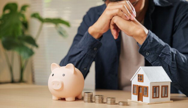 Stressed man holding his forehead, surrounded by a piggy bank, coin stacks, and a house model. Captures the struggles of money issues, financial risks, property concerns
