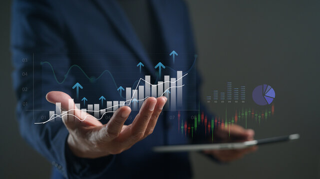 A businessperson in a suit holds a tablet displaying digital financial graphs and charts with rising arrows, symbolizing growth and data analytics.