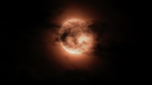 Full red moon rising behind dark clouds in starry night sky.