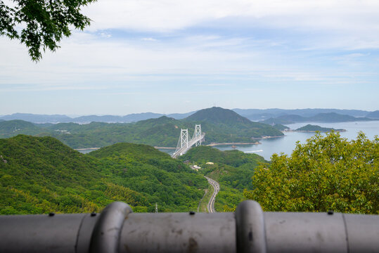 Scenic landscape view of a suspension bridge crossing the sea with lush green mountains and islands in the Seto Inland Sea, framed by foreground elements