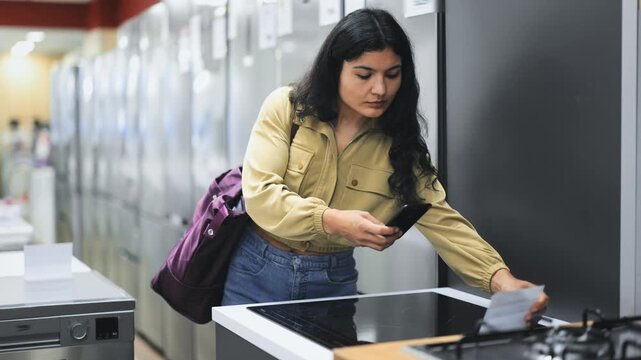 Attentive young woman scanning barcode on ceramic hob while shopping inside consumer electronics market. High quality 4k footage